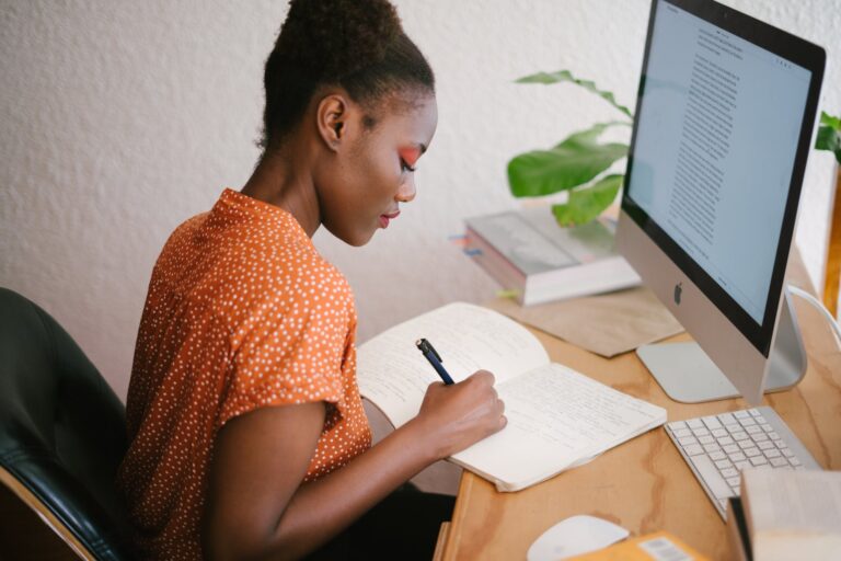 Woman in orange dress writing in notebook whilst sat in front of computer screen