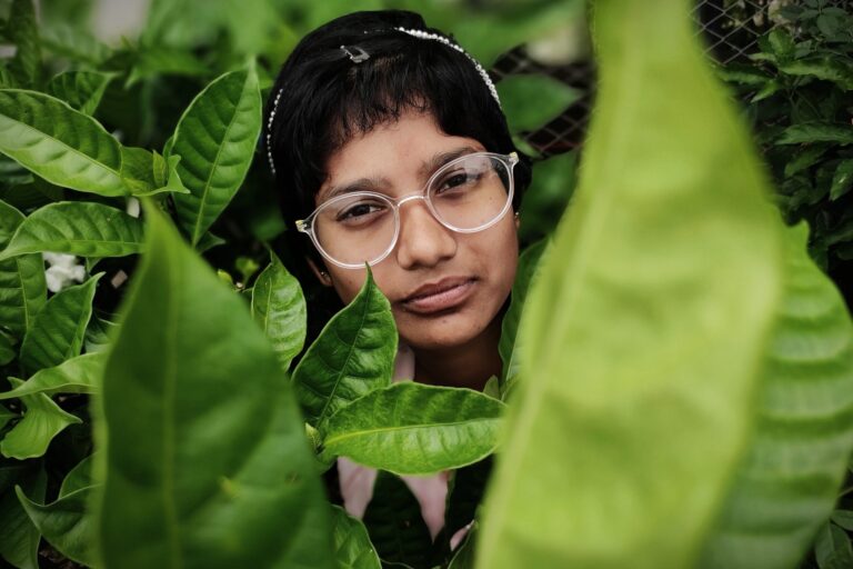 Brunette woman wearing clear framed glasses whilst stood amongst a forest of green leaves