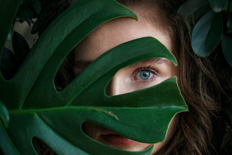 Brunette woman with blue eyes looking through monstera plant leaves