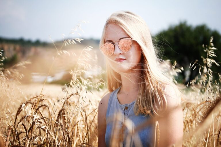 Blonde woman stood in a sunny wheat field wearing reflective rose gold sunglasses